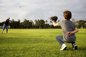 A dad throws a baseball to his kid to catch at the park.