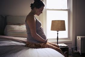 An image of a pregnant woman sitting on a bed at home.