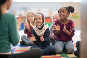 An adorable multi ethnic preschool children are singing and clapping their hands as their teacher is seated in front of them.