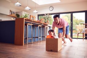 Father And Son Pushing Son Around Kitchen Floor At Home In Junk Modelled Car