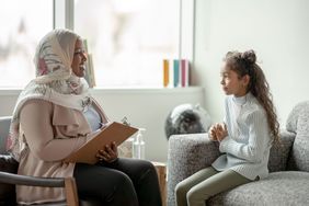 Young girl sits on a couch talking to a therapist holding a clipboard and wearing a hijab