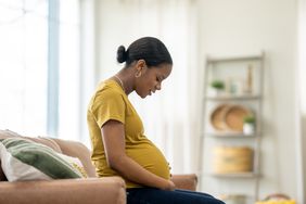 A young pregnant person sits on a sofa, cradling their belly with their hands.