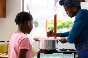 Older Black woman stirs a pot on a gas stove as a young girl watches her. 