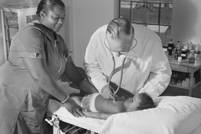 In a black and white photo, a Black nurse helps a white doctor examine a Black child in 1940. 