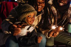 A Black toddler in a hat smirks as a smiling man and woman hold sparklers. 