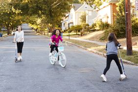 Three girls riding on scooters and a bike in the street