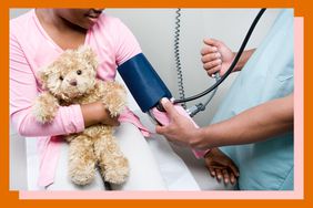 Doctor checking a girl's blood pressure while she holds a teddy bear.