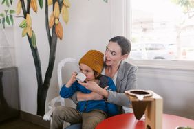 Young woman carrying her little child wearing beanie drinking from little cup sitting at coffee and chocolate boutique shop 