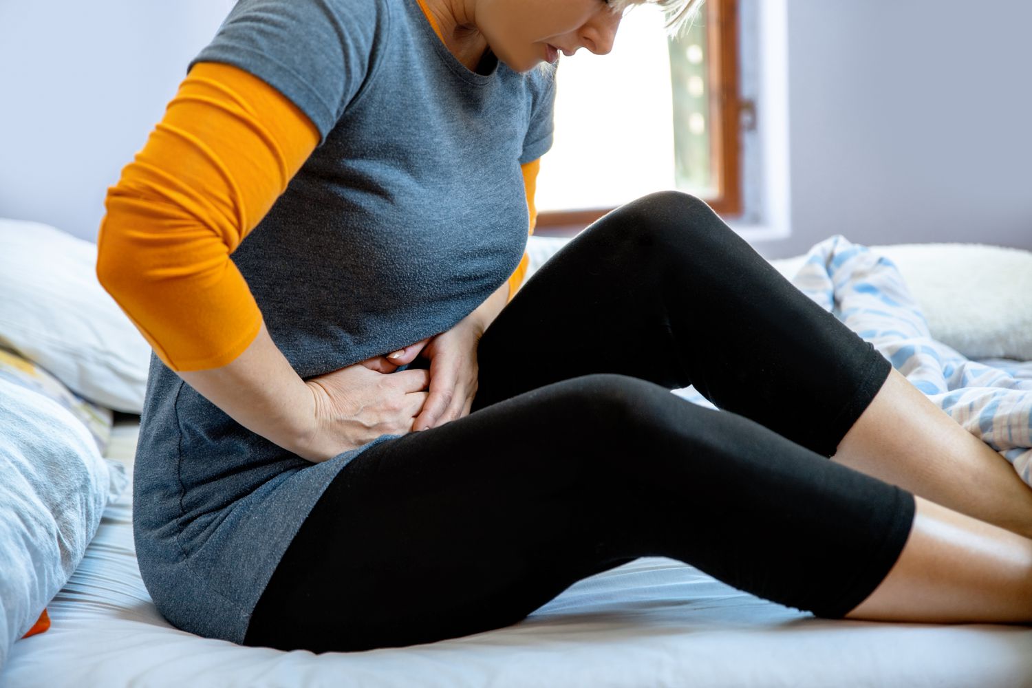 Woman presses her stomach in pain, sitting on bed