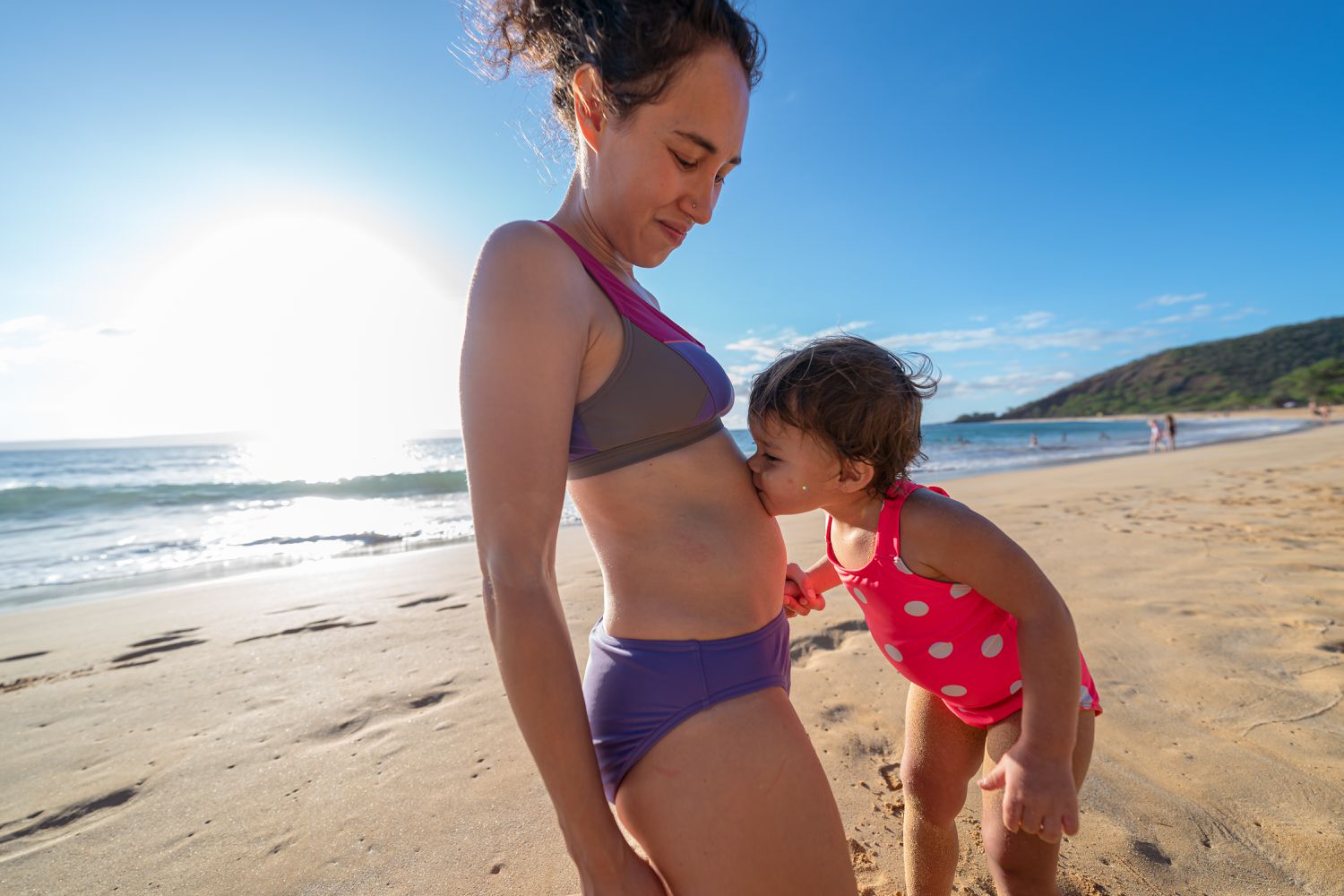 A young woman who is pregnant in the first trimester kneels in the sand as her adorable toddler daughter kisses her pregnant belly while enjoying a fun and relaxing family trip to the beach.