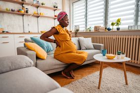 Pregnant person suffering from backache sitting on sofa and holding hands on back and stomach.