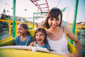 Excited little boy and girl having fun with mother riding amusement park roller coaster