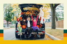 Two children, dressed in Halloween costumes, throw leaves while sitting in the open trunk of an SUV.