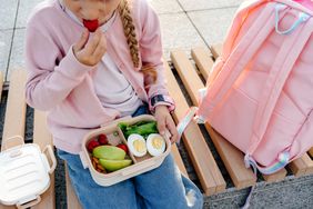 A child eats a strawberry from a lunch box while sitting on a bench