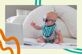 Baby sitting on bed with a laptop wearing sunglasses and a bib.