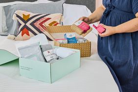 Woman standing opening postpartum recovery kit boxes sitting on bed