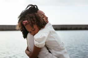 Side view of black mother and daughter gently embracing with closed eyes on blurred background of seascape